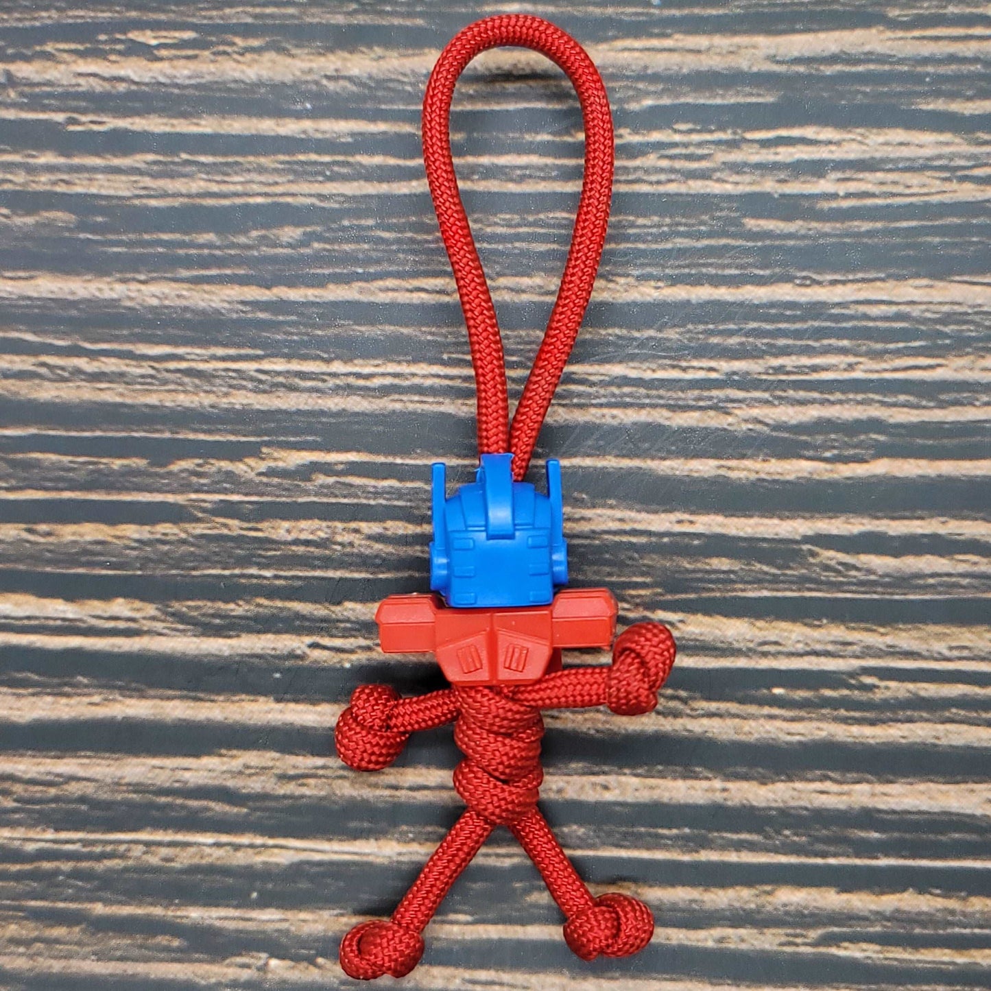 An Insanely Paracord Car Transformer Semi - 550 Paracord Pal Keychain | Handmade | USA-Made | Red | Mini Figure featuring a small, cartoonish figure made from red 550 paracord and plastic parts. The paracord forms the body, legs, and arms, while the plastic parts forming the head give it a Car Transformer semi-robot-like appearance. It lies on a textured wooden surface.