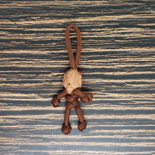 The Insanely Paracord Magic Castle Paracord Pal Keychain - Handmade 550 Paracord Keychain by Abigail & Noah - Cali Brown - High Strength - USA Made is a hand-crafted keychain made of brown 550 paracord, fashioned into the shape of a small humanoid figure with a beige head. The background is a wooden surface with dark and light striped patterns.