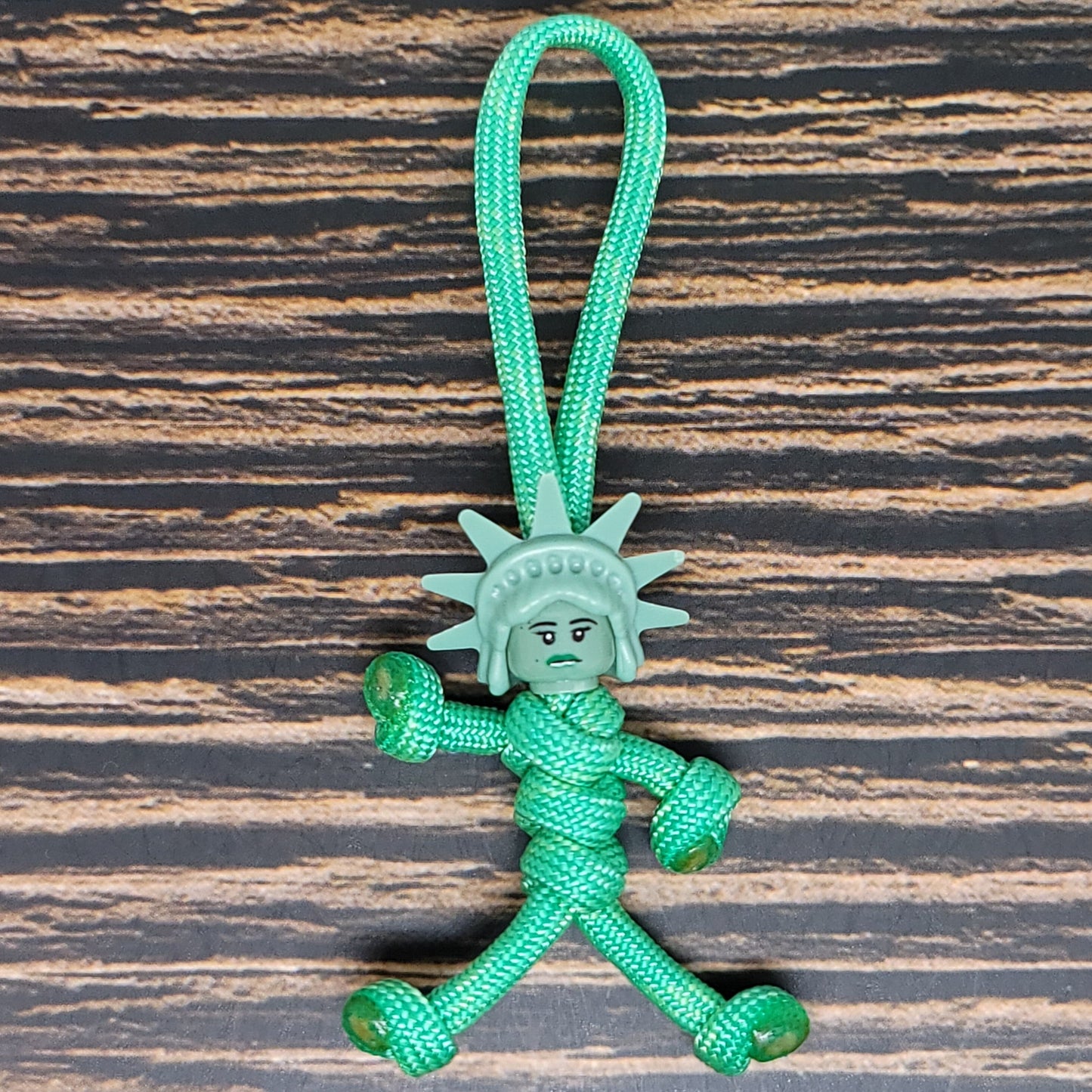 A durable NYC Statue of Liberty Paracord Pal Keychain 550 Cord Strength 2.5 Feet Mint, this small figurine made of green 550 paracord features a face and crown resembling the iconic statue. With arms, legs, and a loop for hanging, it rests on a dark, textured wooden surface.