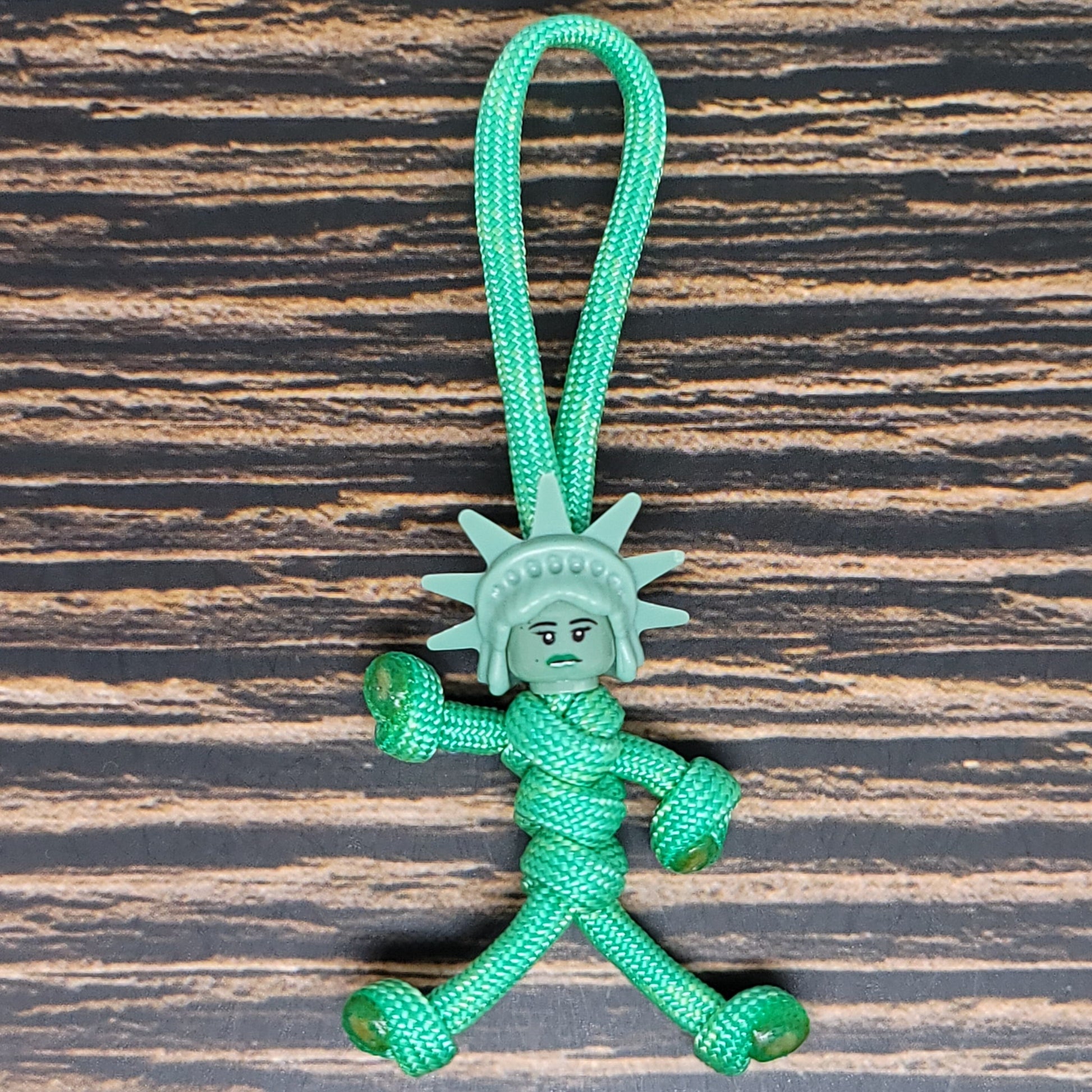 A durable NYC Statue of Liberty Paracord Pal Keychain 550 Cord Strength 2.5 Feet Mint, this small figurine made of green 550 paracord features a face and crown resembling the iconic statue. With arms, legs, and a loop for hanging, it rests on a dark, textured wooden surface.