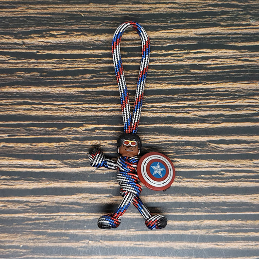 A 550 Paracord Keychain figure wrapped in red, white, and blue cord, resembling a superhero with a shield featuring a blue star in a red circle. The Insanely Paracord Air Force Captain America Paracord Keychain - Handmade with 2.5ft 550 Paracord has a loop at the top for attaching to keys or bags, and the figure has a smiling face with aviator goggles.