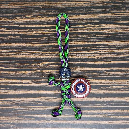 A stylish keychain made of green and purple 550 paracord forms a stick-figure person. The figure has a black helmet, a small shield resembling Captain America's, and is displayed against a wooden surface. Perfectly combining utility and design, this Zombie Shield Captain Keychain - Handmade 550 Paracord Pal adds charm to your accessories. This is an exquisite product by Insanely Paracord.