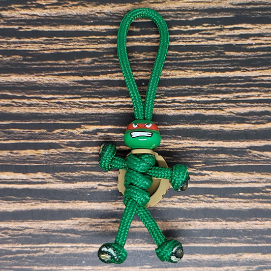 An Insanely Paracord Red Mutant Sewer Turtle Keychain featuring a small, green, rope figure resembling a Teenage Mutant Ninja Turtle with a red mask is placed on a wooden surface. The figure has a loop at the top for attaching to keys or bags, making it perfect for turtle lovers seeking a unique design.