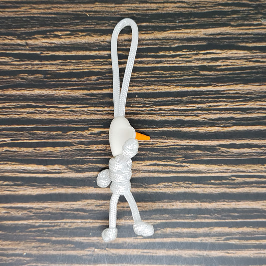 A Magic Castle Snowman - Handmade Paracord Pal Keychain with 550lb Strength - USA Made - Cali White - Unique High Quality Design crafted from light gray 550 paracord, shaped like a small figure with limbs, a white bead for a head, and an orange nose. Dubbed the Insanely Paracord Magic Castle Snowman keychain, it sits against a dark wood surface with light brown grain patterns.