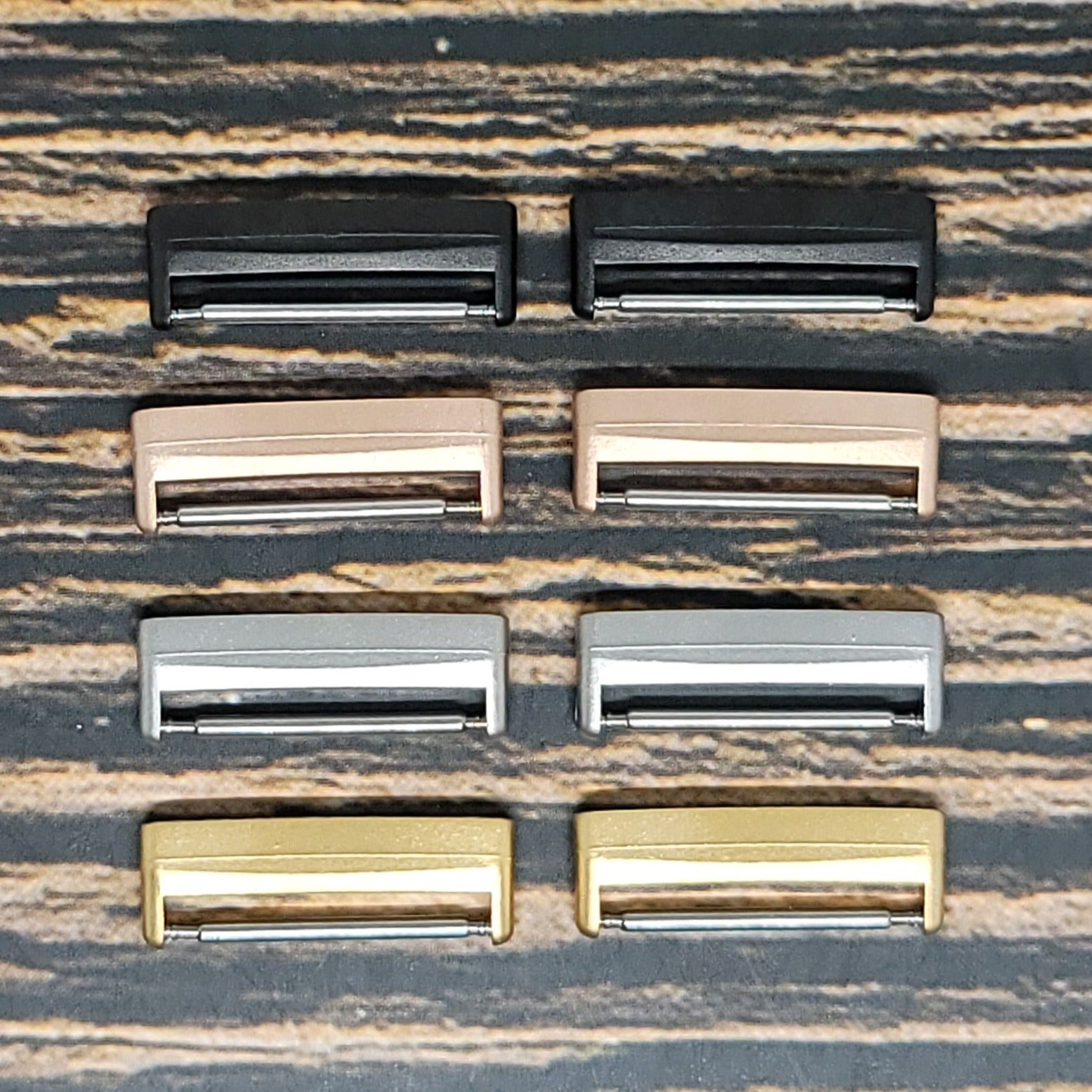A set of eight metal belt buckles arranged in two rows, each in different colors: black, rose gold, silver, and gold. The buckles are placed on a textured surface with brown and black stripes resembling Insanely Paracord's Fitbit Versa 3 Watch Band Adapters - 2 Smart Watch Adapters Included for personalized Fitbit Versa 3 bands.