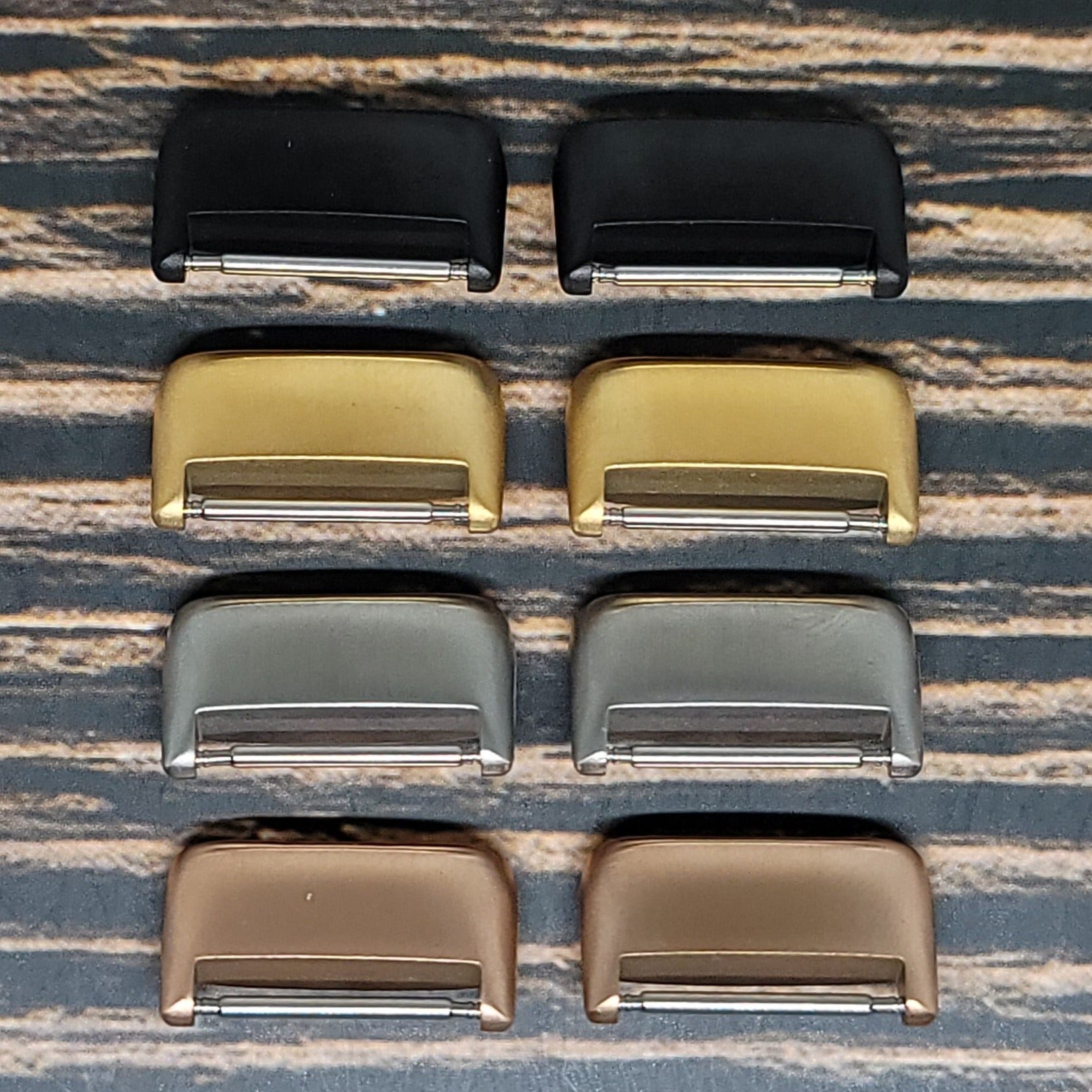 Eight Insanely Paracord Fitbit Charge 5 Watch Band Adapters, resembling guitar bridges, are arranged in two parallel rows on a textured wooden surface. The adapters come in various metallic finishes: black, gold, silver, and bronze. Each row contains four adapters placed next to each other.