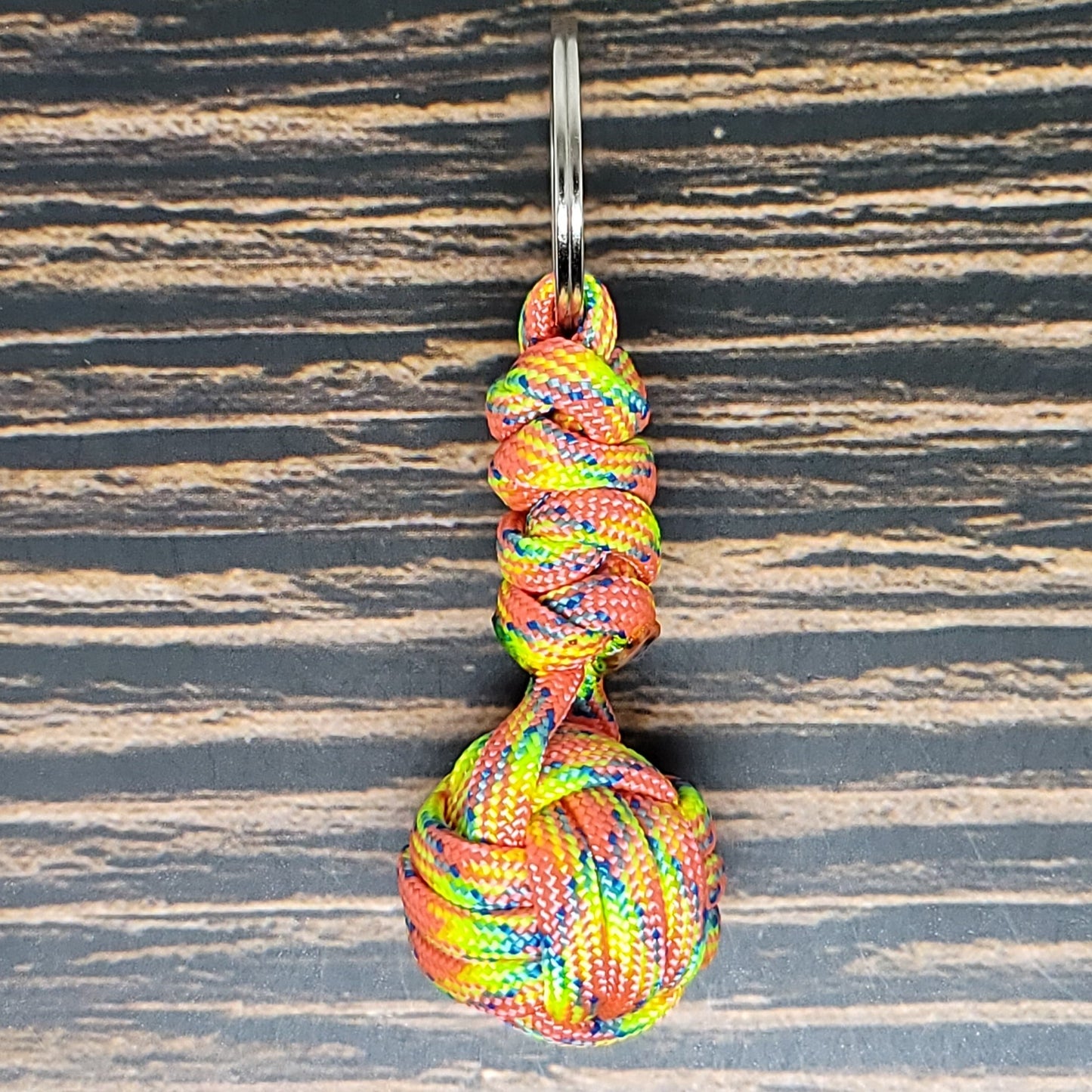 A colorful Monkey Fist Paracord Key Fob with 550 Paracord, 1" Key Ring, and 3/8" Marble lies on a wooden surface, made from multicolored 550 paracord featuring hues of red, yellow, green, and blue. It forms a spherical shape at one end and is topped with a metal ring for attachment, doubling as both a stylish accessory and survival tool. The product is from the brand Insanely Paracord.