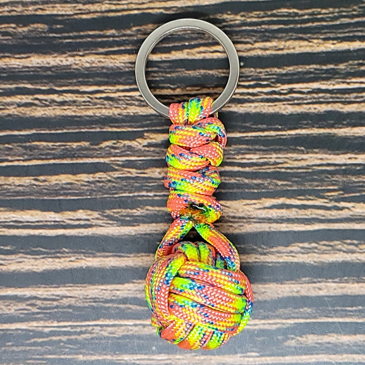 A colorful Insanely Paracord Monkey Fist Paracord Key Fob with 550 Paracord, 1" Key Ring, and 3/8" Marble with a knotted ball and loop design, attached to a metallic keyring, is placed on a wooden surface with dark and light brown stripes. The 550 paracord features a mix of red, blue, yellow, and green hues. This versatile keychain also doubles as a handy survival tool.
