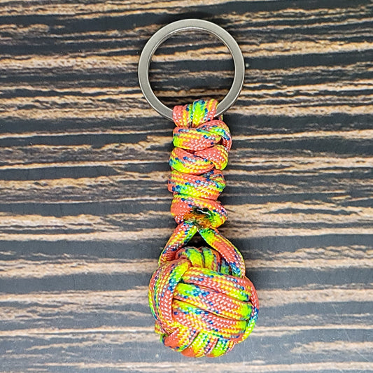 A colorful Insanely Paracord Monkey Fist Paracord Key Fob with 550 Paracord, 1" Key Ring, and 3/8" Marble with a knotted ball and loop design, attached to a metallic keyring, is placed on a wooden surface with dark and light brown stripes. The 550 paracord features a mix of red, blue, yellow, and green hues. This versatile keychain also doubles as a handy survival tool.