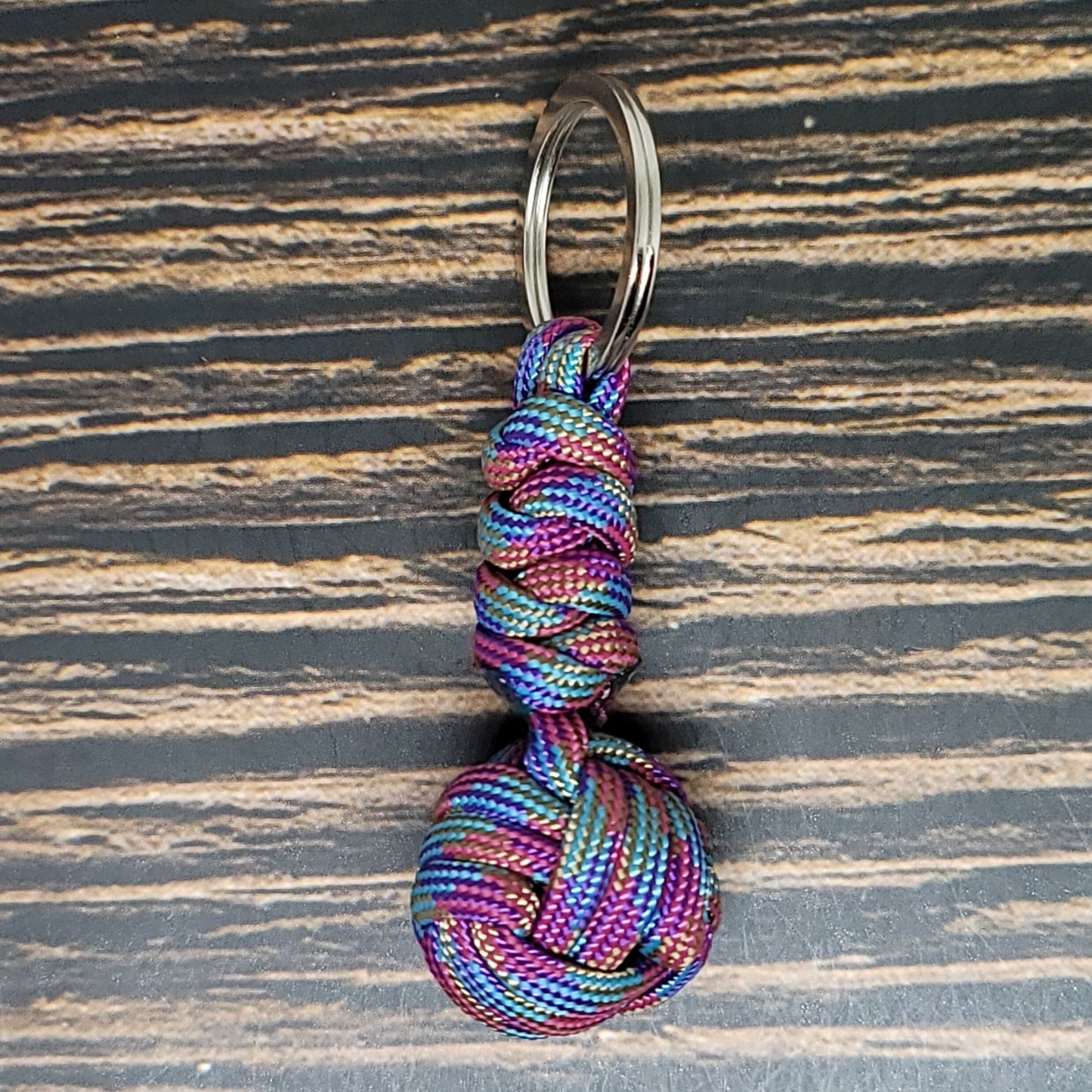 An Insanely Paracord Monkey Fist Paracord Key Fob with 550 Paracord, 1" Key Ring, and 3/8" Marble, crafted with vibrant rainbow colors and intricately tied into a monkey fist knot, sits elegantly on a dark wooden surface. The keyring is securely attached at the top of this stylish survival tool.