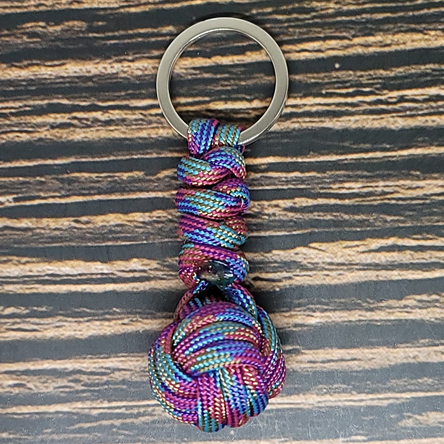 An Insanely Paracord Monkey Fist Paracord Key Fob with 550 Paracord, 1" Key Ring, and 3/8" Marble featuring a colorful knot attached to a metal keyring. Woven with 550 Paracord in shades of purple, blue, green, and pink, this compact survival tool is displayed on a wooden surface with dark and light streaks.