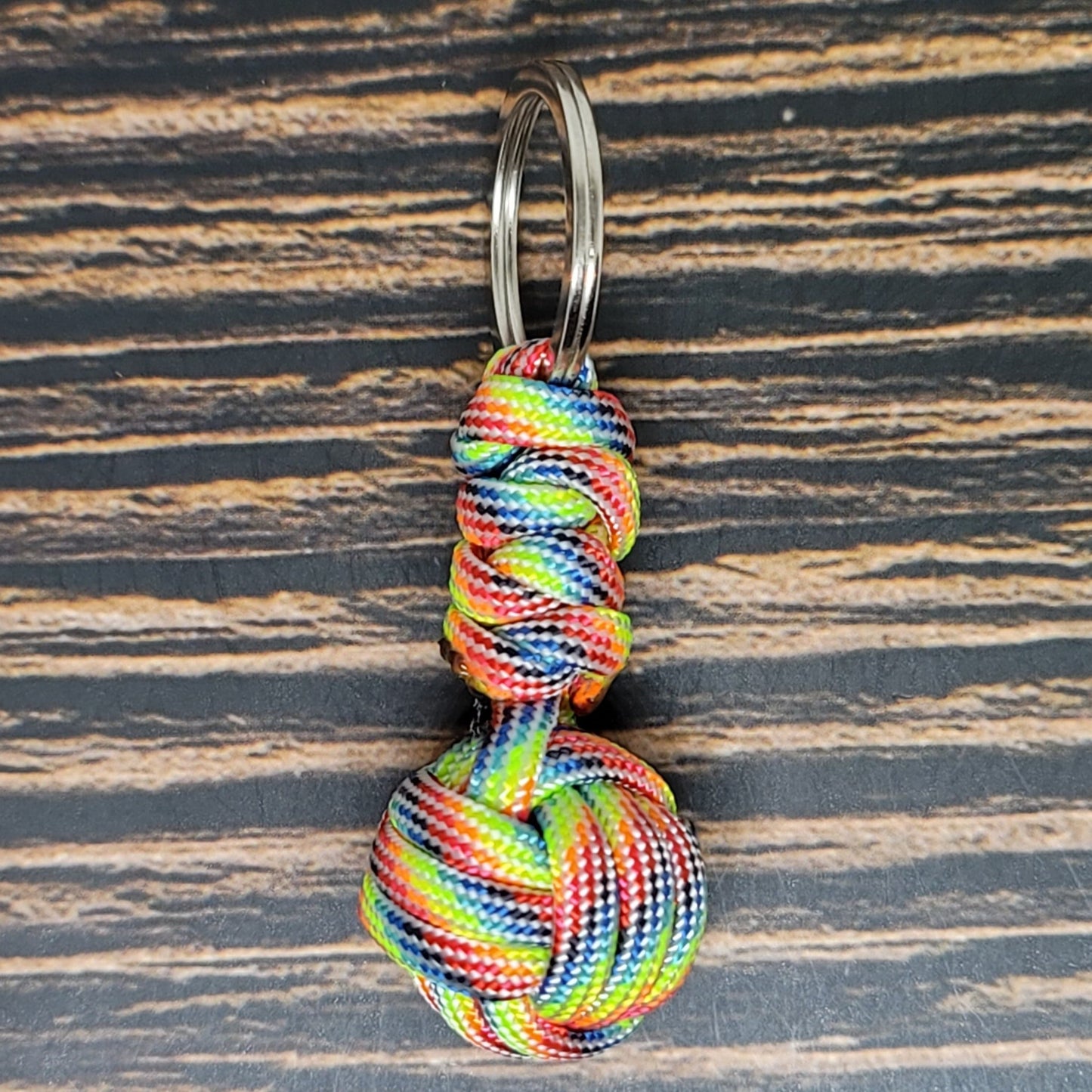 An Insanely Paracord Monkey Fist Paracord Key Fob with 550 Paracord, 1" Key Ring, and 3/8" Marble made from multicolored 550 paracord showcases a skillfully woven spherical shape, complete with a loop at the top attached to a metal keyring. This unique piece not only serves as an eye-catching accessory but also doubles as a handy survival tool. The background features a natural wooden grain pattern.