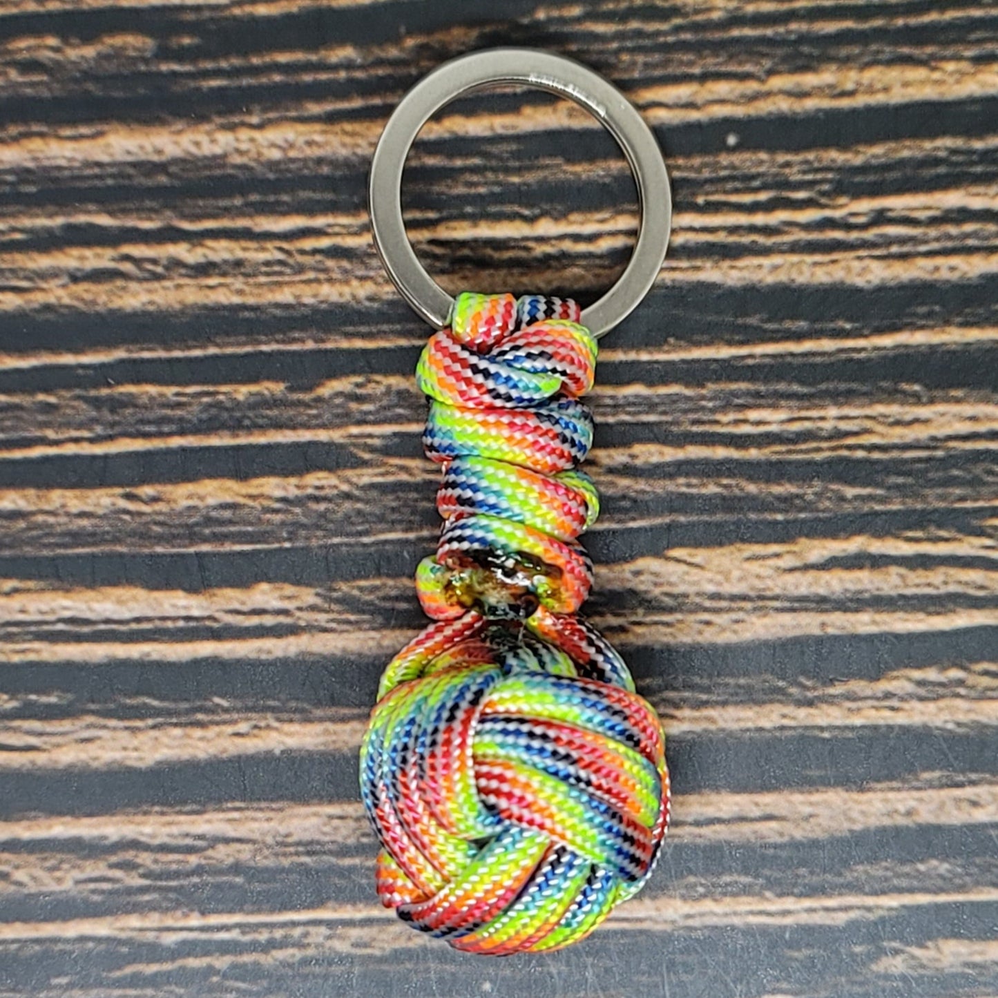 A keychain with a silver ring attached to a multicolored, intricately knotted 550 Paracord forming a decorative pattern sits on a wooden surface with dark and light brown streaks. The Insanely Paracord Monkey Fist Paracord Key Fob with 550 Paracord, 1" Key Ring, and 3/8" Marble features a vibrant mix of red, blue, green, yellow, and orange colors.