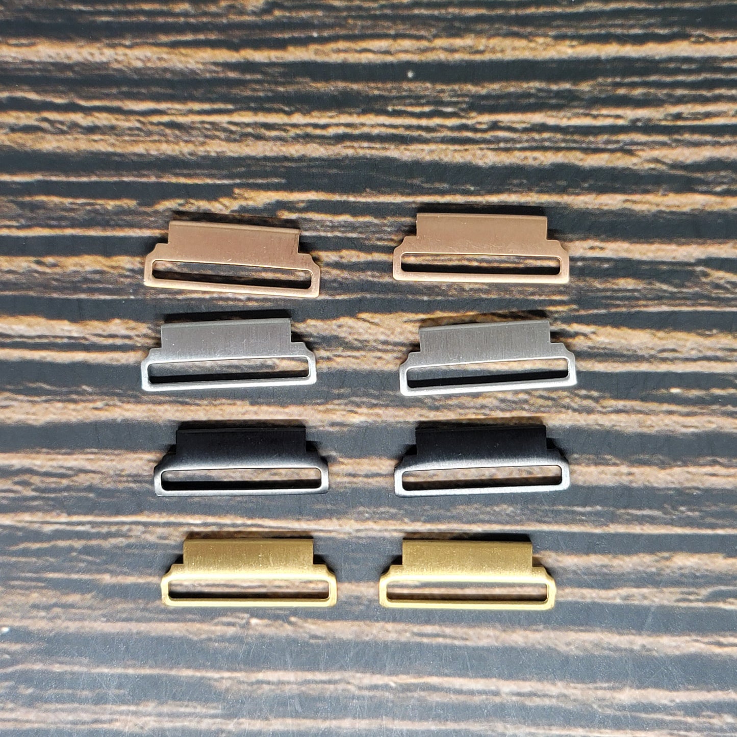 A set of six rectangular metal belt buckles arranged in two rows of three on a wooden surface. The buckles, perfect for integrating with Insanely Paracord's Smart Watch Band Adapter for Fitbit, Huawei, Samsung - 18mm 20mm 22mm Sizes, come in different colors: black, silver, bronze, and gold. Each buckle has a flat frame with a small horizontal bar across the center.