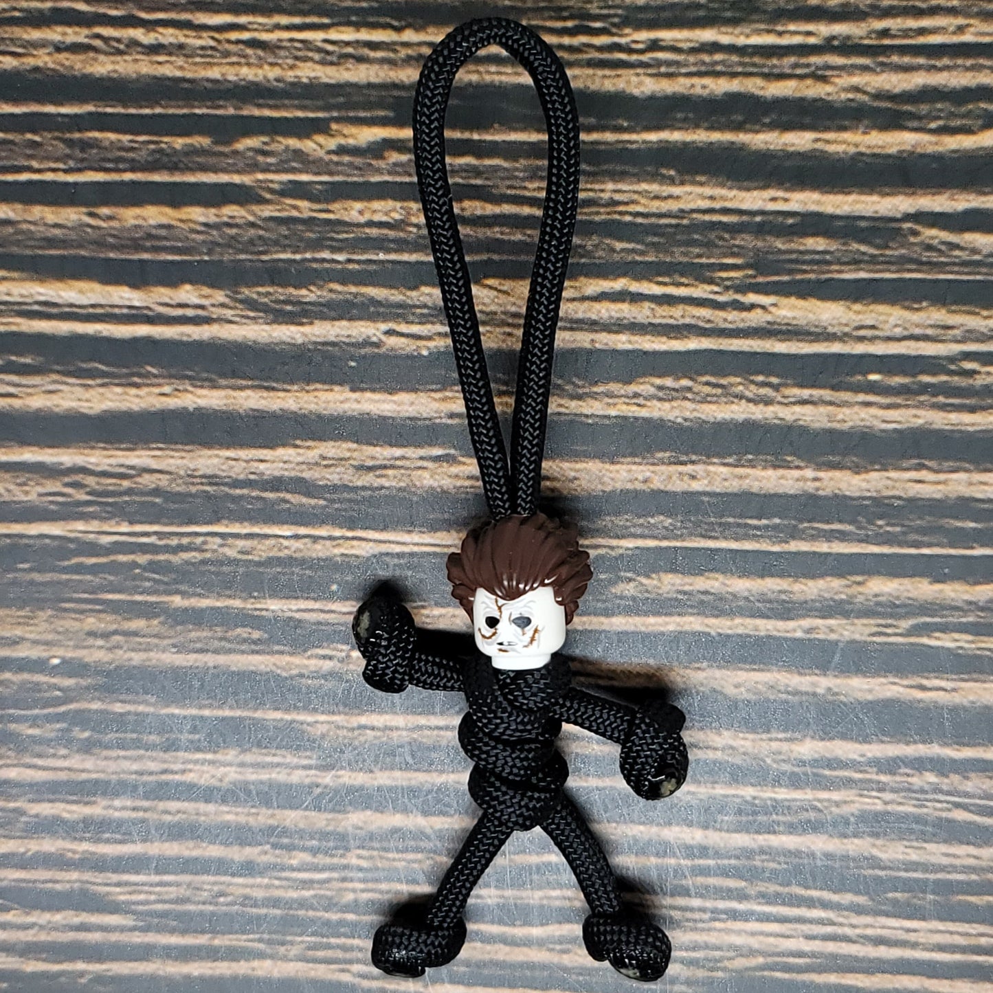 A small figurine with a rough, cartoonish face and spiky brown hair attached to a black paracord rope sits against a wooden surface. This Mike Scary Movie Serial Killer Halloween Paracord Pal Keychain by Insanely Paracord figure is tied to resemble a stick person, making it an eerie and playful accessory for horror movie fans.