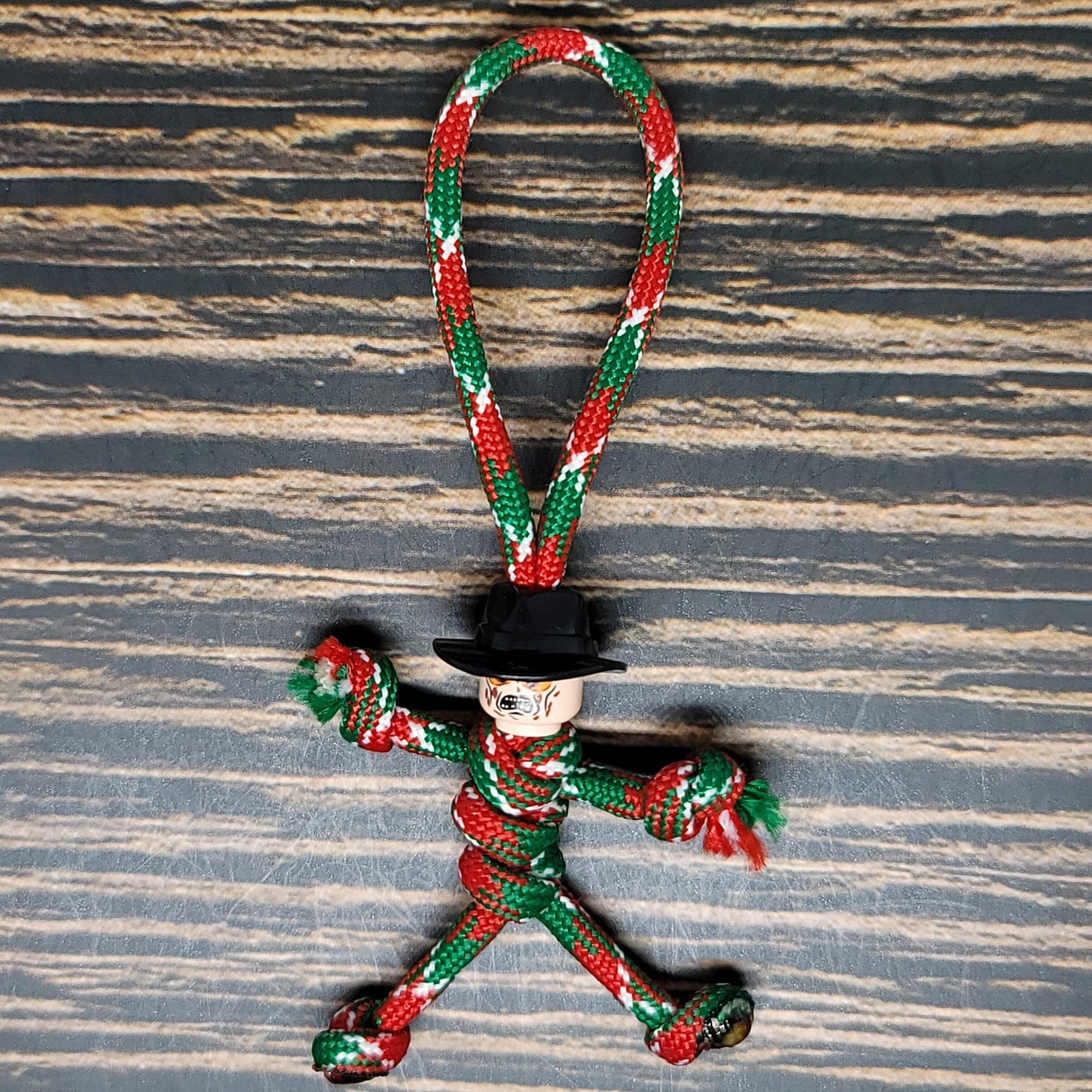 A playful red, green, and white rope toy in the shape of a person with knotted limbs. Crafted from 550 Paracord, it sports a small black hat on its head and is placed on a textured wooden surface. This is the Scary Serial Killer Paracord Keychain by Insanely Paracord.