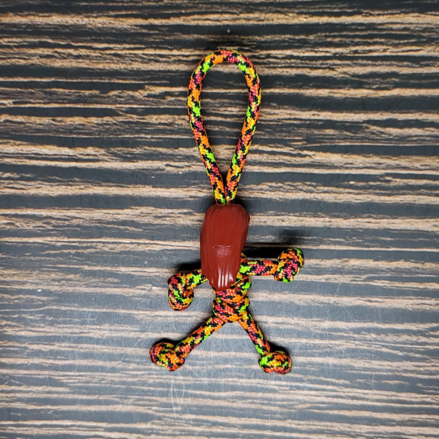 A colorful Nightmare Pumpkin Queen Keychain | Handmade Paracord Pal, shaped like a stick figure person with a brown wooden bead as the head, lies on a striped wooden surface. This handmade Halloween accessory from Insanely Paracord features vibrant colors including green, yellow, orange, and pink, making it a festive and unique addition to any collection.