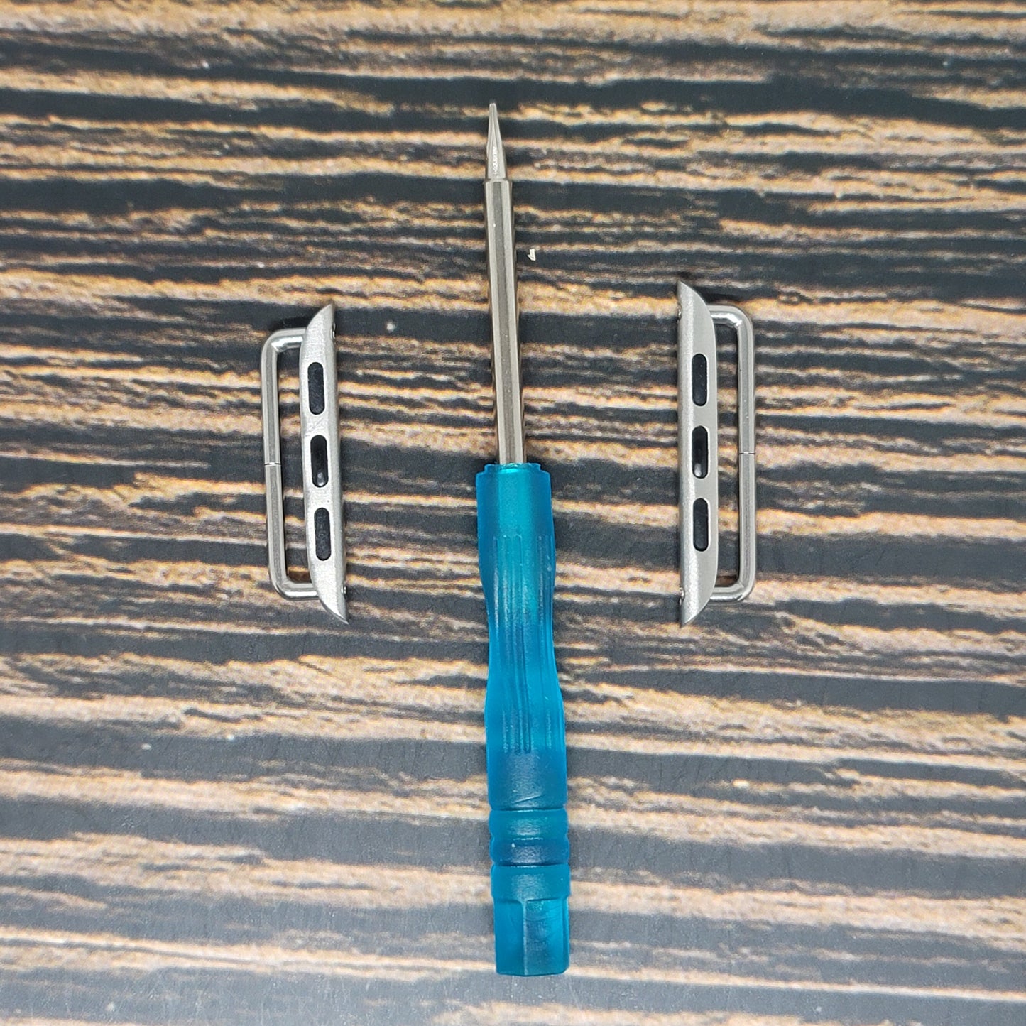 A small blue-handled screwdriver with a Phillips head is placed between two rectangular metal brackets on a wood-grain surface. The brackets, resembling Insanely Paracord Smart Watch Band Adapter for Fitbit, Huawei, Samsung - 18mm 20mm 22mm Sizes, have three holes each, suggesting they are mounting brackets or clips for attaching something.