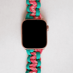 A smartwatch with a square face and black screen is attached to an Insanely Paracord Custom Paracord Watch Band - Durable, Stylish, Made in USA. The band features intertwined pink and turquoise strands, making it both colorful and durable. This smartwatch-compatible accessory rests on a plain, light-colored surface.