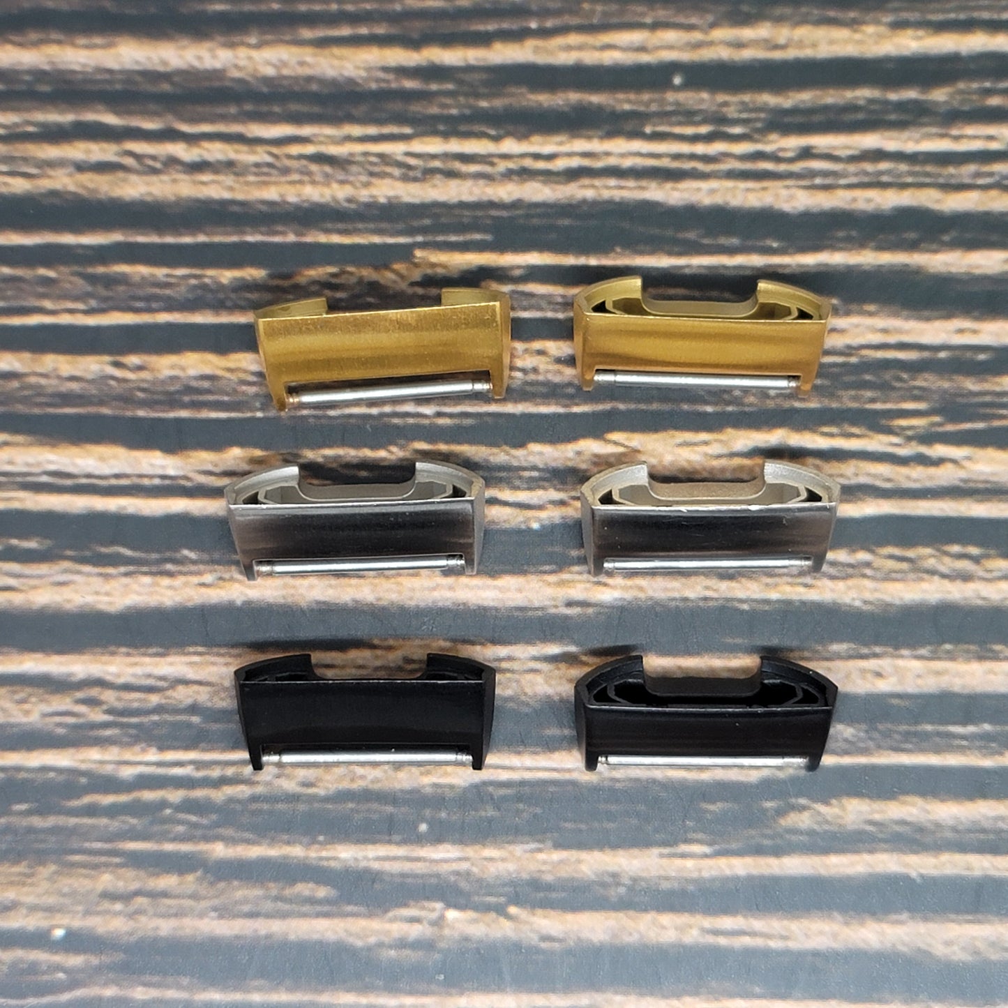 A set of six metal guitar saddles, arranged in two rows of three, shares the stage with a fine Insanely Paracord Fitbit Watch Band Adapter - Convert Your Smartwatch with Adaptors in Four Colors. The top row features gold, silver, and chrome saddles while the bottom showcases black, nickel, and black nickel on a textured wooden surface. Perfect for any compatible Fitbit models.