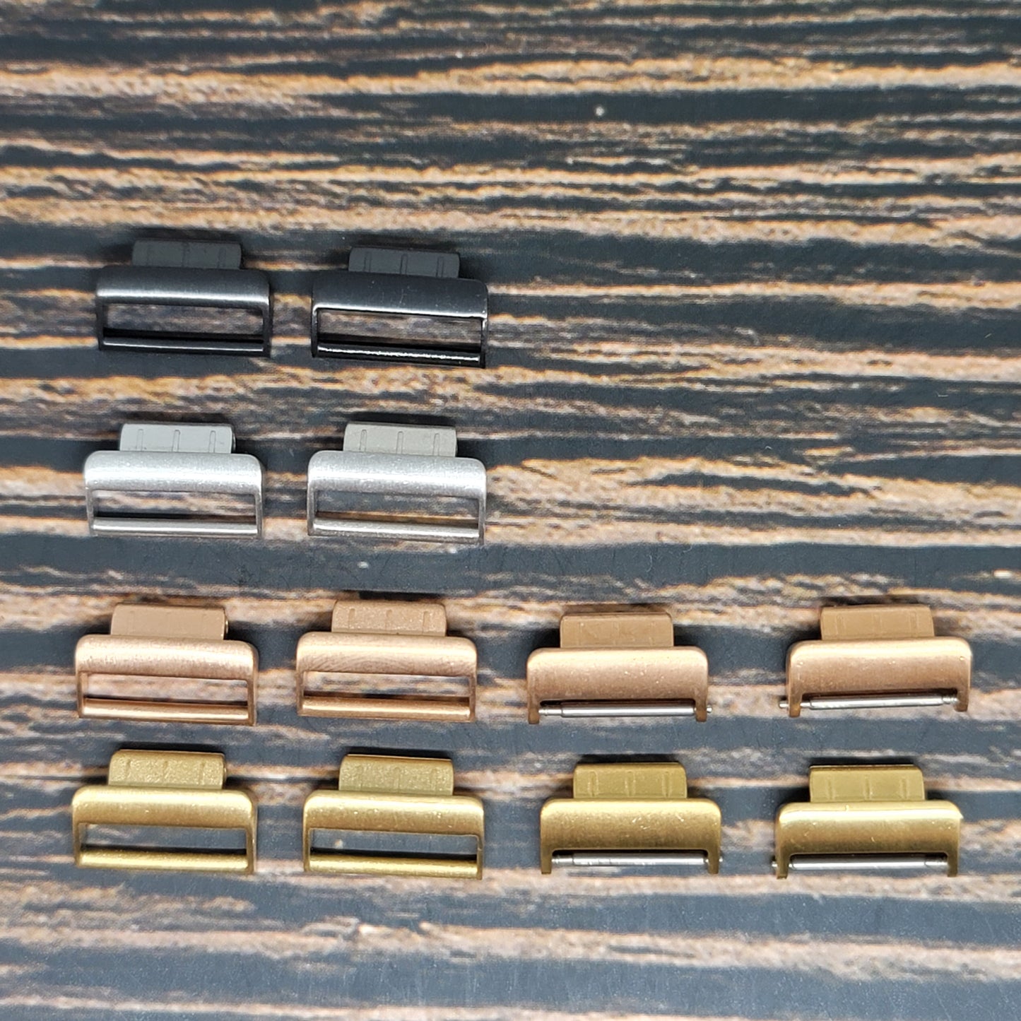 A collection of variously colored Fitbit Watch Band Adapter - Convert Your Smartwatch with Adaptors in Four Colors, compatible with Fitbit models, displayed on a wood-textured surface. The adapters are organized in a grid pattern with three rows and five columns, showcasing different finishes including black, silver, bronze, and gold.