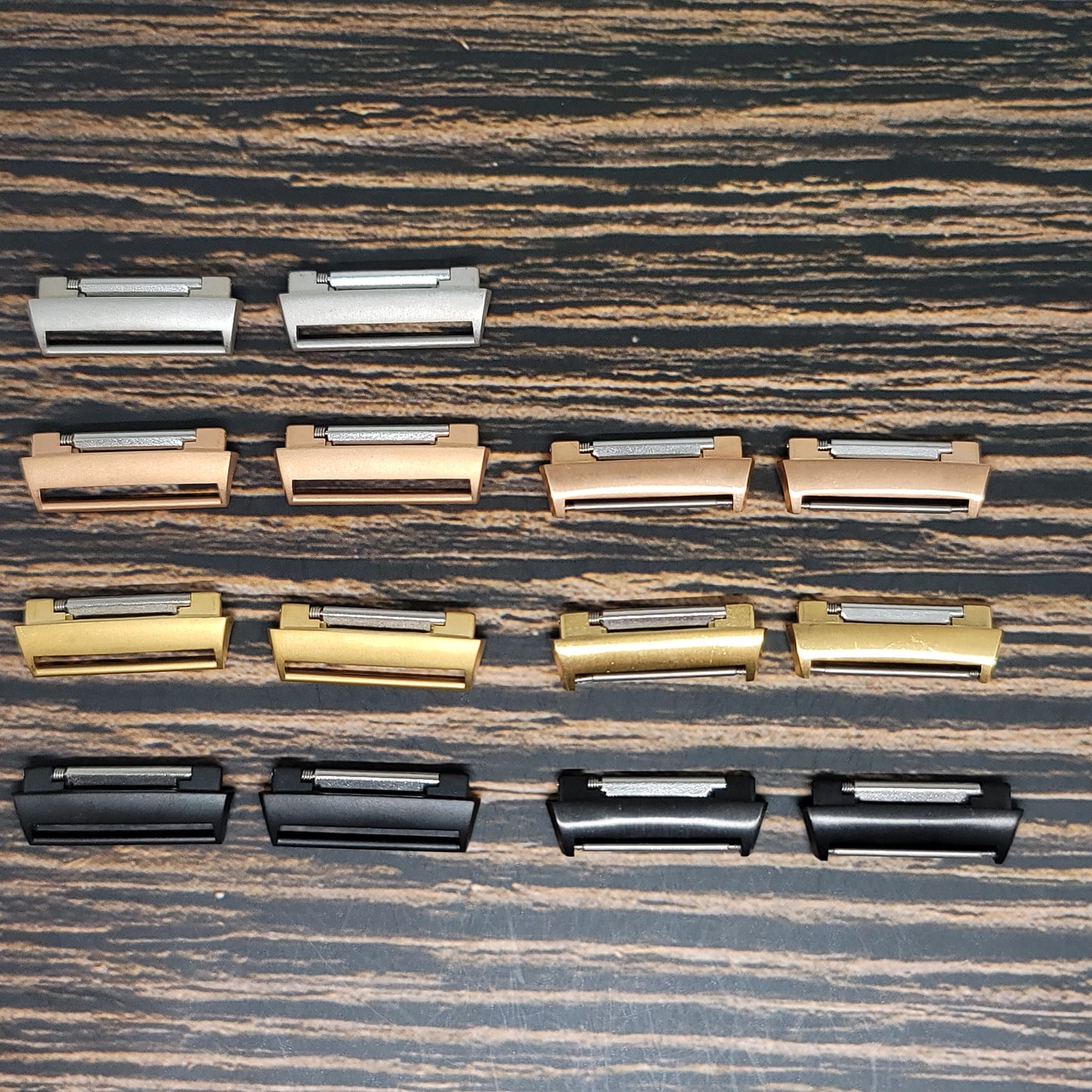 An array of Fitbit Watch Band Adapter - Convert Your Smartwatch with Adaptors in Four Colors by Insanely Paracord organized in four rows on a wooden surface. From top to bottom, the rows feature silver, bronze, gold, and black adaptors respectively. Each row contains four identical adaptors aligned horizontally, much like a neatly arranged smartwatch style display.