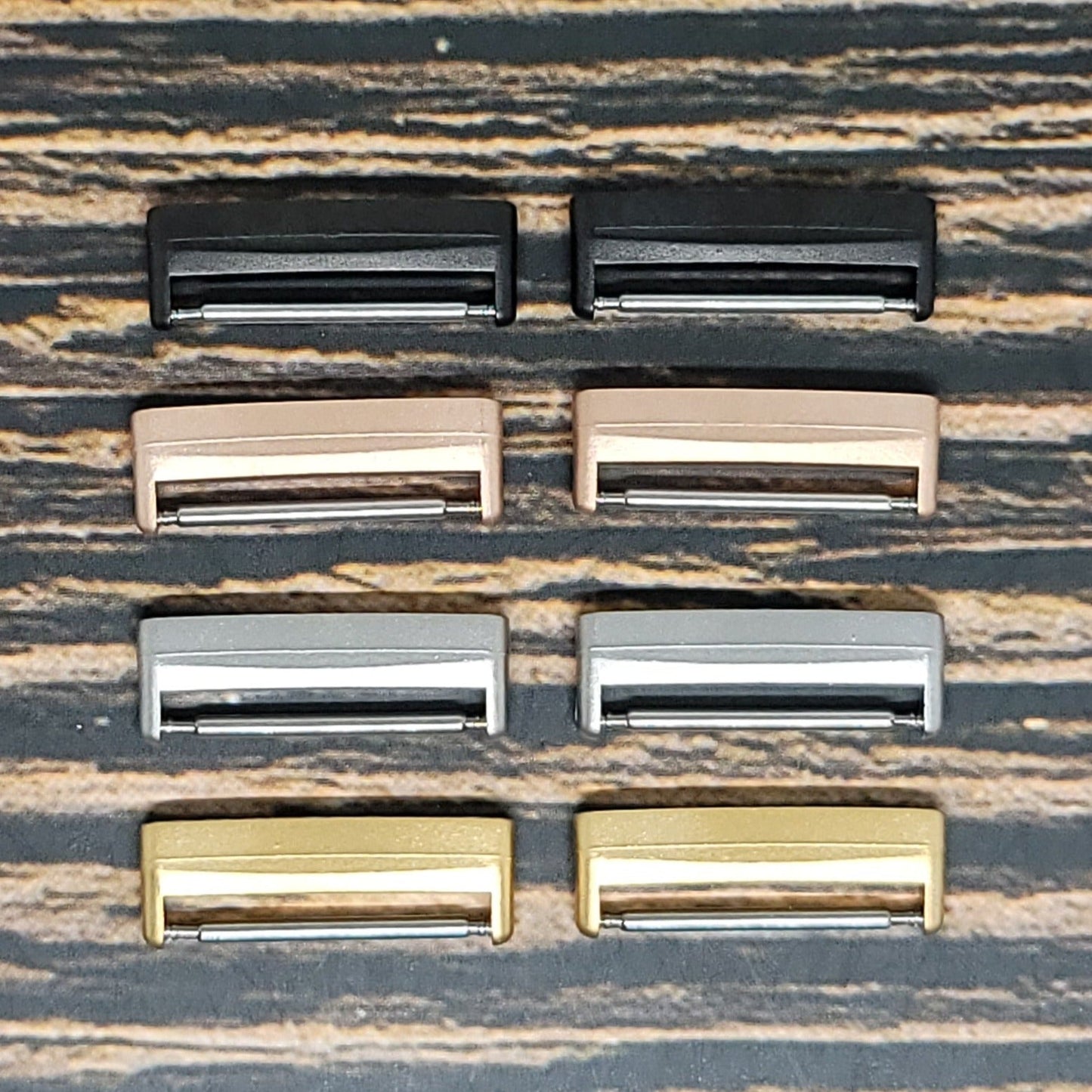 A set of eight rectangular belt buckles in different metallic finishes—two in black, two in rose gold, two in silver, and two in gold—are neatly arranged on a striped wooden surface. These buckles feature a rectangular frame and bar for securing the belt, making them ideal for use as Insanely Paracord Fitbit Watch Band Adapter - Convert Your Smartwatch with Adaptors in Four Colors.