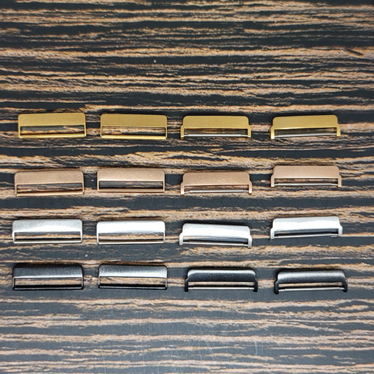 A variety of Fitbit Versa Watch Band Adapters - Create Your Own Custom Fitbit Watch Bands by Insanely Paracord, perfect for custom Fitbit watch bands, are arranged in four horizontal rows on a wooden surface. The adapters come in a variety of colors including gold, bronze, silver, black, and metallic finishes, evenly spread out in an organized manner.