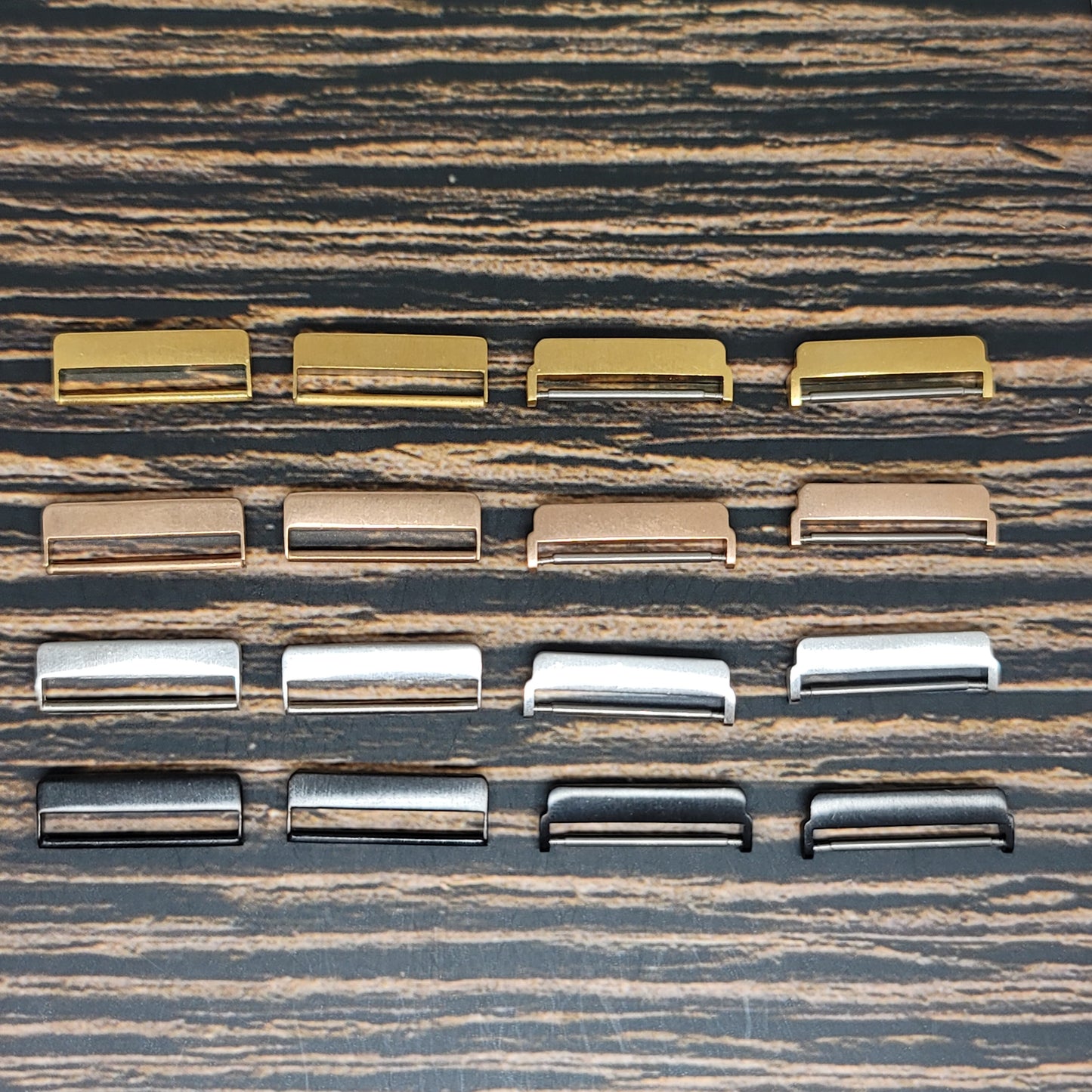 A collection of twelve Fitbit Watch Band Adapters - Convert Your Smartwatch with Adaptors in Four Colors by Insanely Paracord is neatly arranged in three rows on a textured wooden surface. With finishes in gold, bronze, silver, and black, these adapters exhibit a sleek smartwatch style.