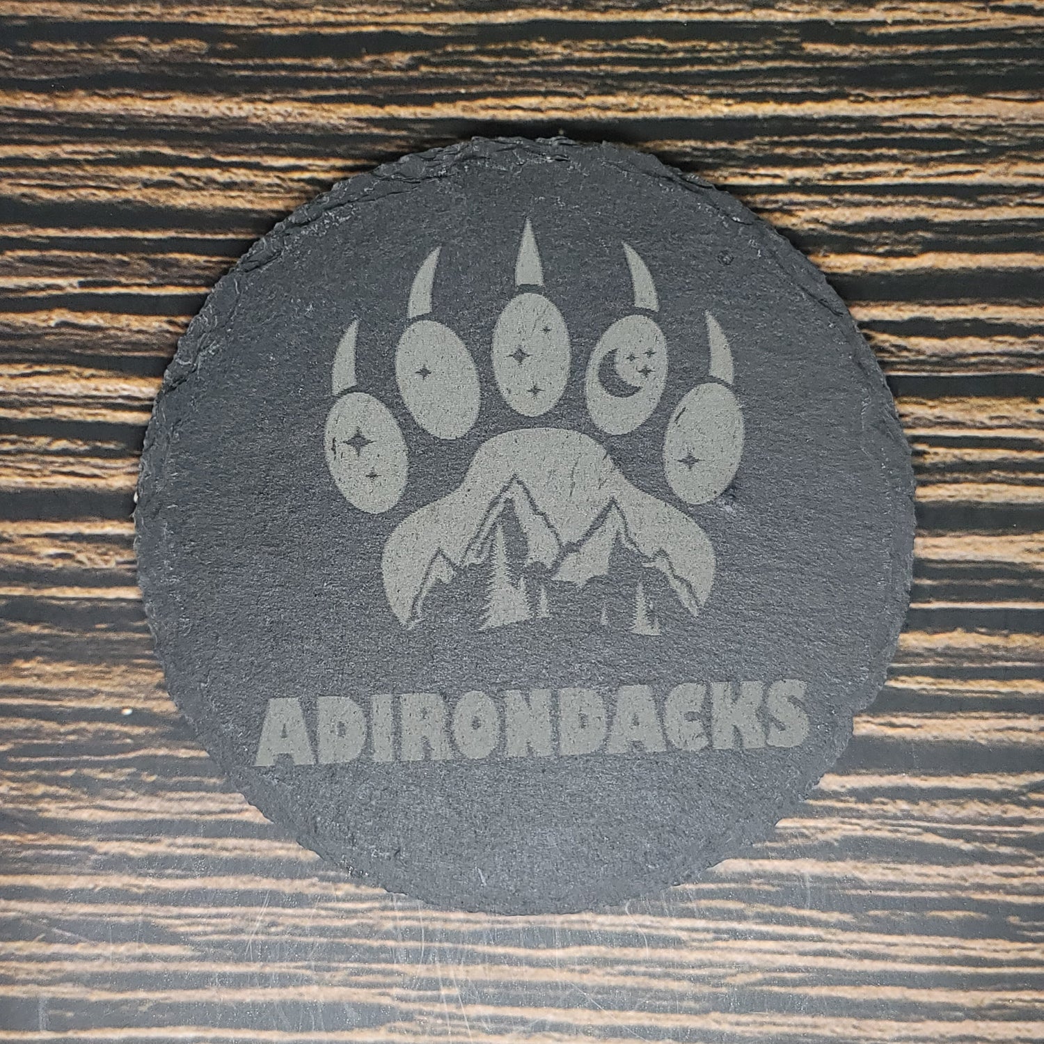 A black oval custom slate coaster with a design featuring a bear paw print, each toe containing a star and moon. Below the paw, there are mountaintops, and underneath the design, the word "ADIRONDACKS" is etched. Perfect as Insanely Paracord Custom Slate 4" Coasters - Personalized Laser Engraved Drink Coasters for a unique corporate gift, it sits on a wooden surface.