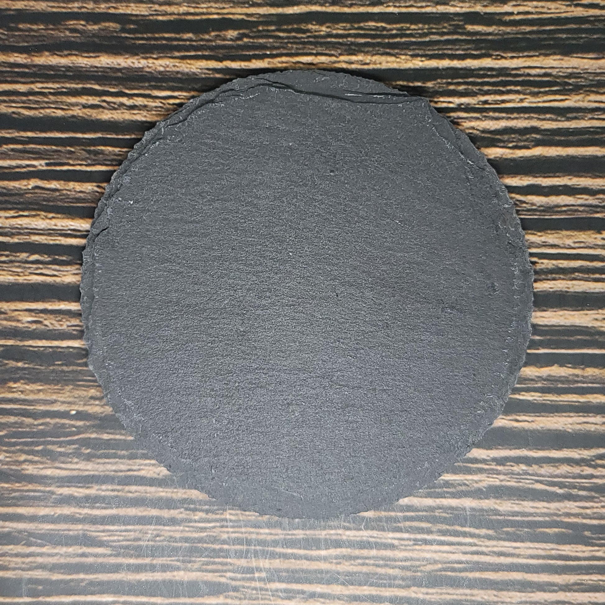 A perfectly round, dark gray Custom Slate 4" Coaster - Personalized Laser Engraved Drink Coaster from Insanely Paracord sits on a table with a wood grain pattern. The coaster has a rough, natural edge contrasting with the smooth surface of the table, making it an ideal corporate gift.