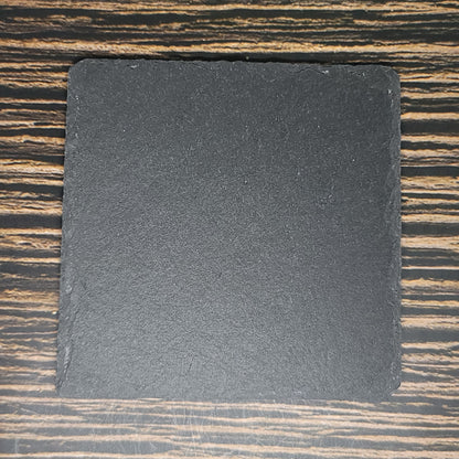 A square black slate tile with rough edges is placed on a wooden surface with a striped pattern. The Custom Slate 4" Coasters - Personalized Laser Engraved Drink Coasters, perfect for custom slate coasters or corporate gifts, is centered in the image and has a matte texture. The wooden surface features a mix of light and dark brown hues. These coasters are brought to you by Insanely Paracord.