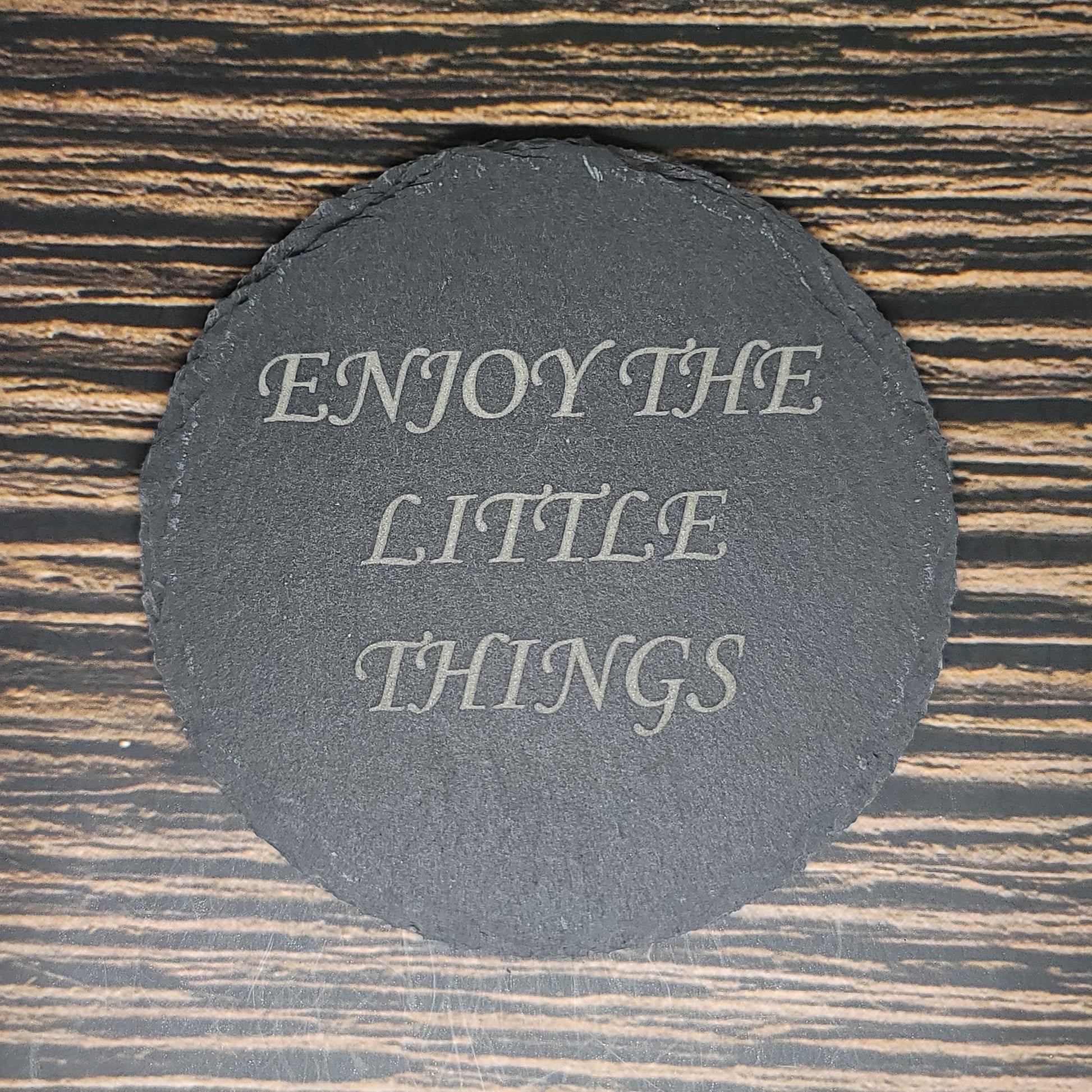 A round, black Custom Slate 4" Coaster from Insanely Paracord with the text "ENJOY THE LITTLE THINGS" written in elegant white letters is placed on a wooden surface with dark and light brown stripes.