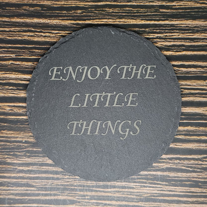 A round, black Custom Slate 4" Coaster from Insanely Paracord with the text "ENJOY THE LITTLE THINGS" written in elegant white letters is placed on a wooden surface with dark and light brown stripes.