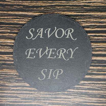 A circular, custom slate coaster with the text "SAVOR EVERY SIP" written in elegant, light-colored script is placed on a wooden surface with horizontal grain patterns. Ideal as a corporate gift, this stylish addition enhances any setting. These Custom Slate 4" Coasters - Personalized Laser Engraved Drink Coasters by Insanely Paracord perfectly blend functionality and aesthetics.