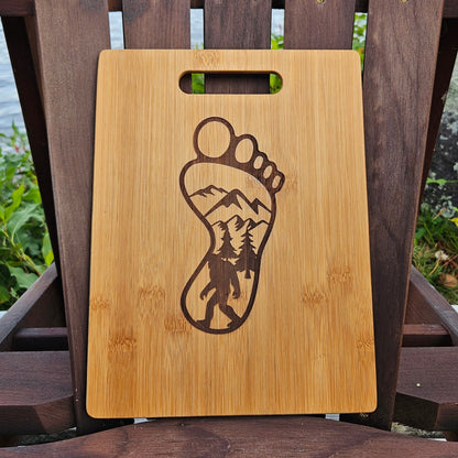 A rectangular wooden cutting board, ideal as a corporate gift, boasts a handle at the top and features a laser-engraved design of a human footprint. Inside the footprint lies a detailed scene of trees and mountains. The Custom Bamboo (12x8) or Acacia (12x9) Cutting Board by Insanely Paracord rests on a wooden chair outdoors, with greenery and water in the background.