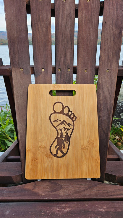 An Insanely Paracord Sacandaga Lake Bamboo (12x8) or Acacia (12x9) Cutting Board with a cutout handle sits on a wooden Adirondack chair. The laser engraved cutting board features a footprint design containing a mountain and forest scene inside. The background reveals a body of water and a mountainous landscape with autumn foliage, ideal for personalized gifts.