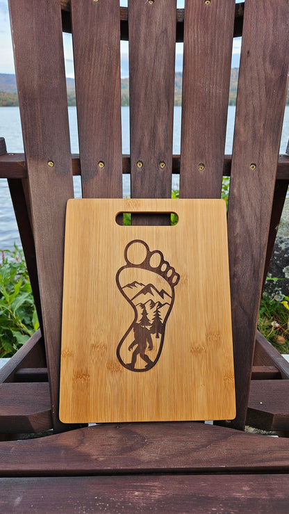 An Insanely Paracord Piseco Lake Bamboo (12x8) or Acacia (12x9) Cutting Board with a handle at the top and a laser engraved footprint design sits on a wooden chair. The footprint features a mountain and forest landscape within its outline. In the background, there is a lake, greenery, and distant hills—an ideal corporate gift.