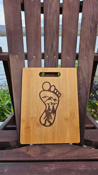 An Insanely Paracord Speculator, NY Bamboo (12x8) or Acacia (12x9) Cutting Board with an engraved design of a human footprint containing a mountain landscape. The custom cutting board rests on a wooden Adirondack chair, with a scenic lake and foliage visible in the background—perfect for those seeking personalized gifts.