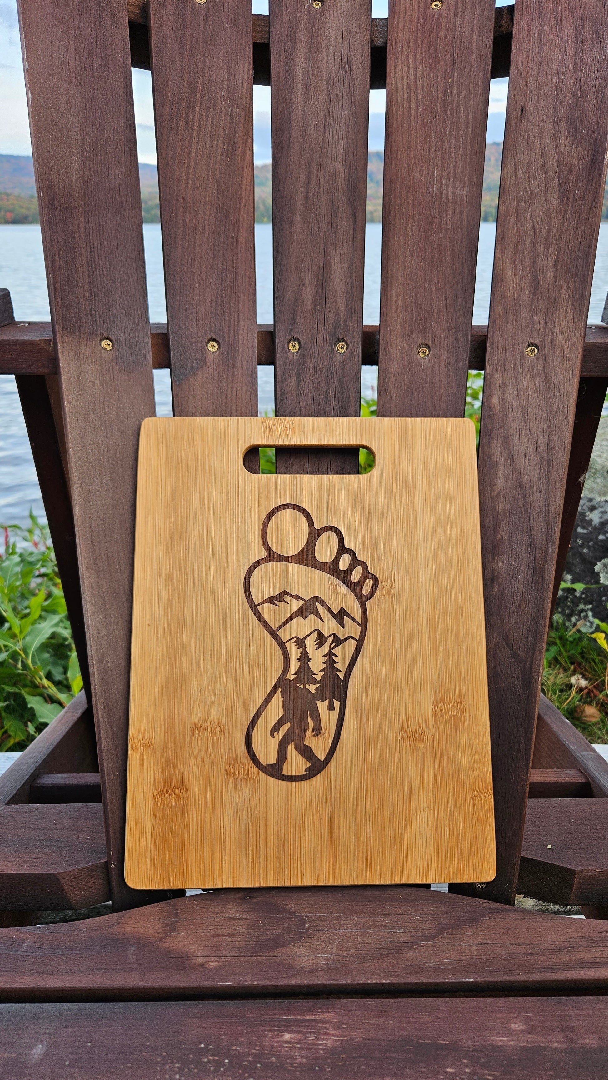 A laser-engraved **Lake Pleasant Bamboo (12x8) or Acacia (12x9) Cutting Board** by **Insanely Paracord** with a footprint design featuring a mountain and forest landscape is placed on a wooden chair. In the background, there is a lake with trees and hills visible on the other side. This custom cutting board makes an ideal corporate gift.