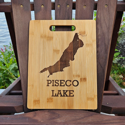 A beautifully laser engraved wooden cutting board, featuring the outline of Piseco Lake and the text "PISECO LAKE," is showcased on an Adirondack chair. The chair sits outdoors, with lush greenery and a serene lake in the background. This custom Insanely Paracord Piseco Lake Bamboo (12x8) or Acacia (12x9) Cutting Board makes for a perfect corporate gift.
