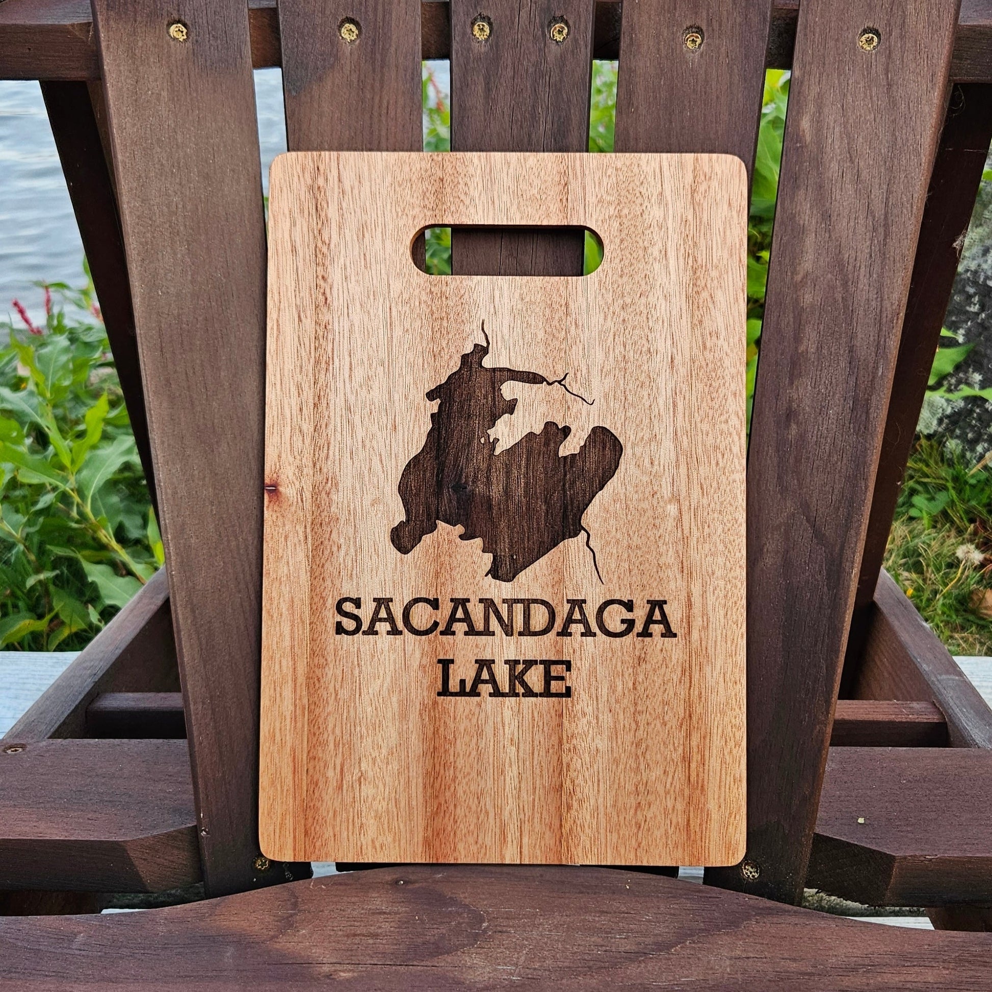 An Insanely Paracord Sacandaga Lake Bamboo (12x8) or Acacia (12x9) Cutting Board is placed on a dark brown wooden chair by a lake. The custom cutting board features a laser engraved image of a lake map with the words "SACANDAGA LAKE" underneath. In the background, there are green plants and water, making it the perfect personalized gift.