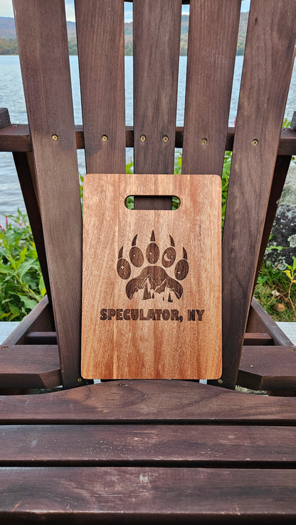 A Custom Bamboo (12x8) or Acacia (12x9) Cutting Board by Insanely Paracord with the text "SPECULATOR, NY" and an engraved design featuring mountains and a bear paw is placed on a wooden Adirondack chair. The laser-engraved piece sits near a lake with hills and foliage in the background, making it an ideal corporate gift.