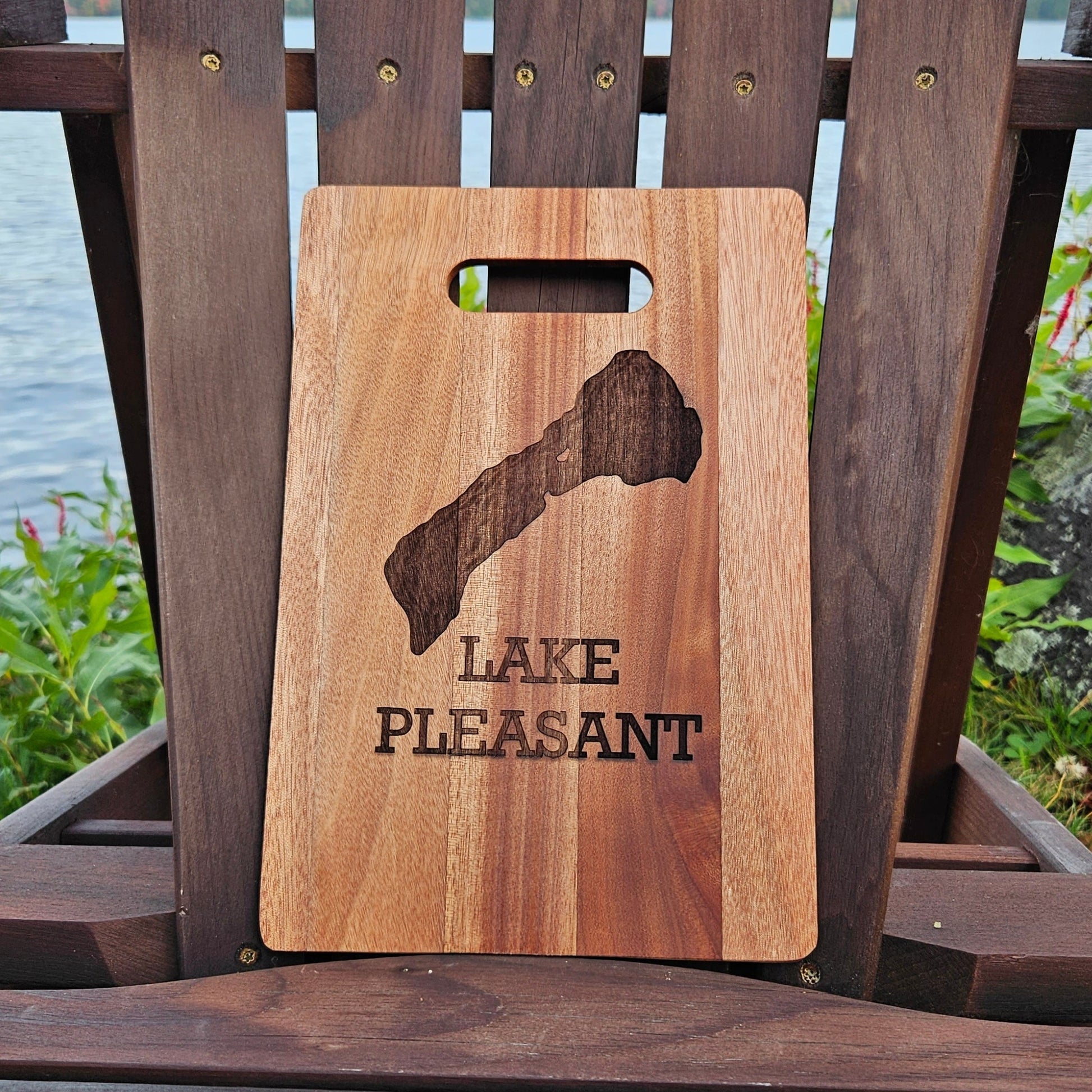 An Insanely Paracord Lake Pleasant Bamboo (12x8) or Acacia (12x9) Cutting Board, laser engraved with an outline of a lake and the words "Lake Pleasant," rests on a wooden chair. With a serene body of water and lush greenery in the background, this custom piece makes an ideal corporate gift.