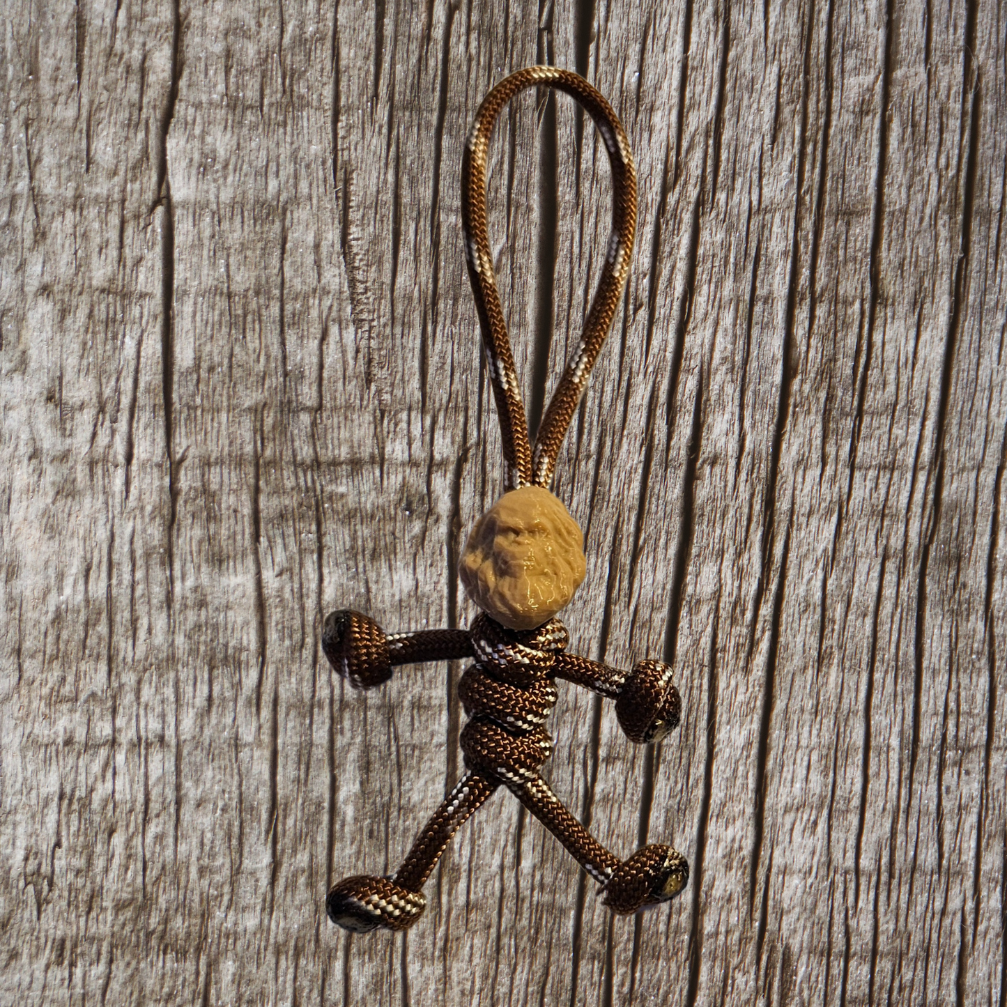 The Insanely Paracord's Bigfoot Paracord Pal Keychain is a handcrafted stick figure made from 2.5 feet of robust 550lb strength paracord and features a meticulously carved wooden head. Made in the USA, this keychain stands out against a textured wooden backdrop, highlighting its distinctive charm and craftsmanship.