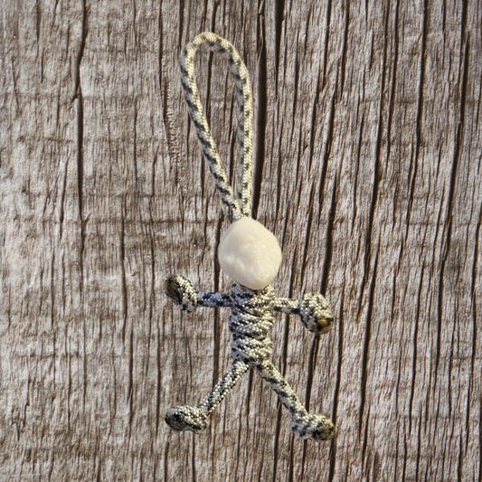 The charming Yeti Paracord Pal Keychain from Insanely Paracord is a handmade figure crafted in the USA using 550 Paracord. It features a white stone for a head and is displayed against a wooden background, showcasing looped rope arms and legs.
