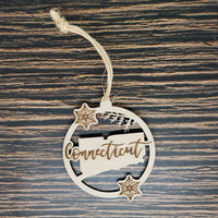 A Connecticut State Christmas Ornament - Laser Engraved Birch Wood from Insanely Paracord featuring the outline of the state of Connecticut with the word "Connecticut" written across it. The custom state ornament hangs from a twine loop and is decorated with two small, intricate snowflakes. The background is a textured wooden surface.