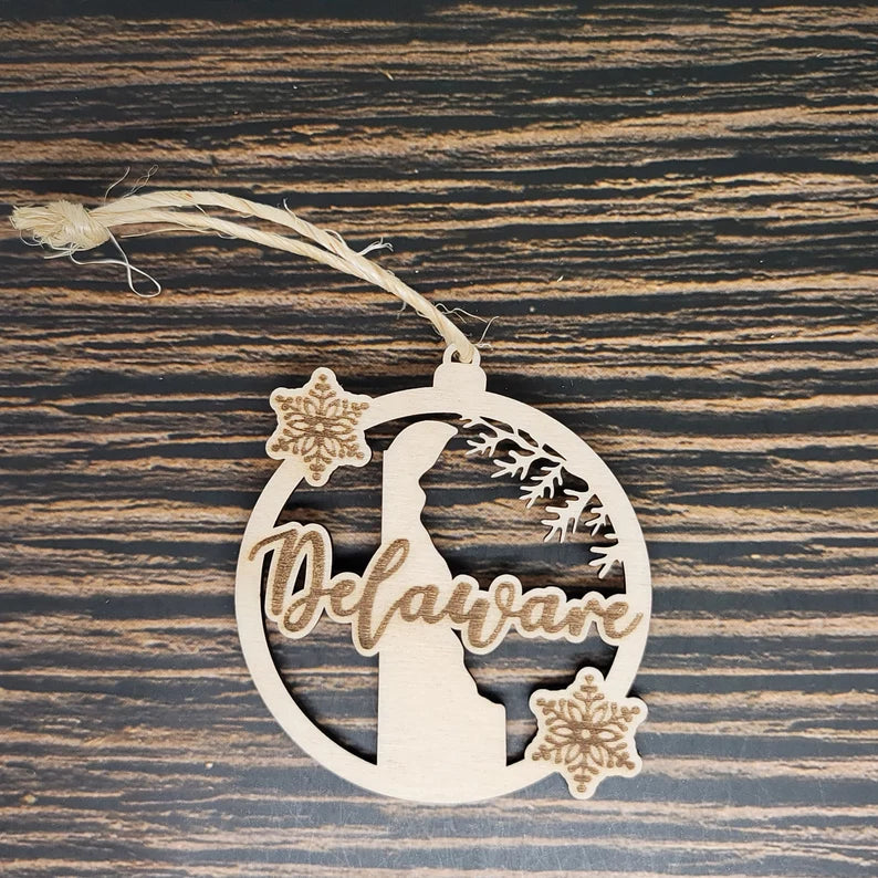 An Insanely Paracord Delaware State Christmas Ornament 3" Laser Engraved Wood featuring the word "Delaware" in script, with the state's outline at the center. Two snowflakes decorate the design, with one at the top right and one at the bottom left. The ornament hangs from a rustic twine loop, perfect for holiday decor.