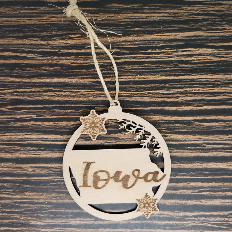 A beautiful Insanely Paracord Iowa State Laser Engraved Christmas Ornament - 3" Tall Birch Wood Holiday Decor, crafted from birch wood, showcases the shape of Iowa with "Iowa" elegantly engraved. This circular holiday decor features snowflakes and pine branches and hangs by twine on a rustic wooden background.