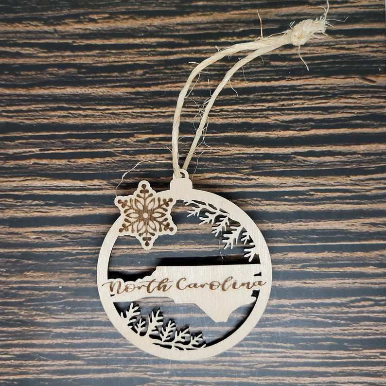 A North Carolina Christmas Ornament - Laser Engraved Birch Wood, 3" Tall from Insanely Paracord lays flat on a dark wood surface. The high-quality birch wood piece is circular and features a laser-engraved snowflake and tree branch design with the shape of North Carolina and the text "North Carolina" engraved in the center. A string is attached at the top.
